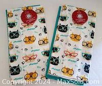 Two folded kitchen towels side by side showing cat face patterns with glasses and small bee prints.