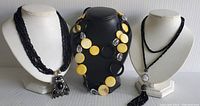Three vintage costume jewelry necklaces on display busts: black multi-strand with rectangular pendant, black and yellow linked with large discs, and black beaded with tassel pendant.
