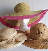 Three sun hats displayed; a Papillon wide-brim straw hat with pink edge, a beige unbranded paper hat with large bow, and a brown unbranded paper hat with woven cutout design.