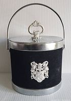 Front view of vintage black ice bucket with silver lid, handle and crest emblem on front