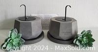 Two new concrete tabletop fire pits seen from an angle with metal hook apparatus on top, gray faceted concrete with black round bases, accompanied by green succulent plants.