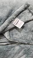 Close-up photo showing the soft plush texture and the sewn edge of the gray throw blanket with a partially visible care label.