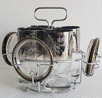 Set of six glass highball glasses with silver floral engravings arranged in metal wire stand with handle, surrounded by six matching round silver floral lusterware coasters mounted on stand sides.