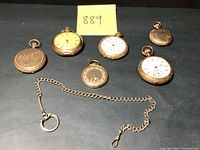 Six pocket watches, one empty case and silver-tone chain laid out on black surface
