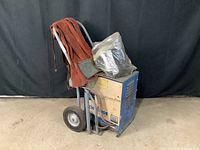 Side view of blue and beige Chloride welding machine on wheeled frame with red leather welding jacket and plastic-wrapped face shield.