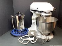 White KitchenAid Classic Plus stand mixer with stainless steel bowl and three attachments in front, Frieling French press on Le Creuset blue enamel trivet behind.