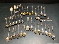 Full set of 35 antique collector spoons laid out showing various sizes and types, mostly silver with some gold tone bowls and ornate handles.