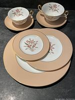 Photo showing a set of tea cups and saucers along with various plates featuring peach borders and floral central designs with gold trim