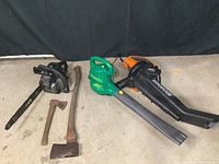 Full view of all 5 yard tools including chainsaw, two axes, and two handheld blowers/vacuums on a concrete floor with black background