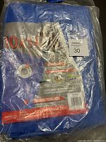 Photo of the folded blue tarp in its original package with visible label showing size 10x14 feet and details about the crossweave material and grommets.