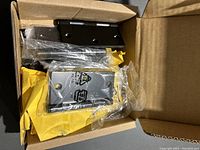 Box showing black square door hinges inside plastic wrap in an open cardboard box.