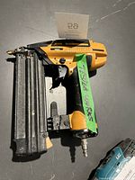 Left side view of a yellow and black Bostitch pneumatic nail gun, showing the metal magazine and air hose connector with identification tape attached.