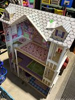 Front and side view of large doll house showing three floors with decorated wallpaper and rugs, white shingled roof, and detailed balcony railings.