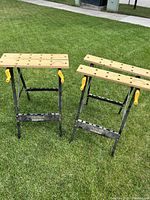 Pair of metal folding work tables with wooden tops and yellow handles on a lawn.