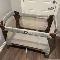 Graco Pack & Play playpen shown from a top angle in a room. Rectangular shape with mesh sides and padded bottom with patterned fabric. Dark brown and gray color scheme.