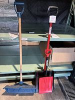 Two snow shovels standing side by side on a shelf, one with a long wooden handle and blue blade, the other red blade with metal handle.