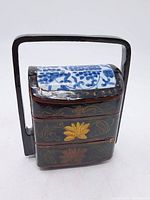 Front view of the 2-tier trinket box showing black wooden body with gold/red floral designs and blue-white floral porcelain tile lid. Black handle attached to the sides.