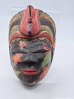 Front view of carved wooden mask with floral painted details and bird carving on forehead.