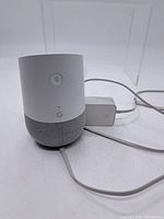 White Google Home smart speaker showing mute button, status LED, and fabric lower half with attached power cable.