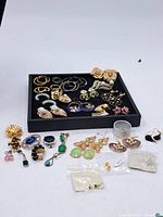 Overview of the lot showing a variety of earrings, brooches, and small jewelry pieces on a black tray and laid out on a white surface