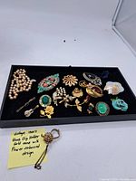 Full display of the assorted vintage jewelry laid in a black tray with a yellow note describing a vintage 1940s glove clip holder in gold tone with flower embossed design.