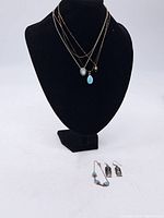 Three gold-tone chain necklaces displayed on a black bust, featuring a blue teardrop pendant, a white stone pendant, and a small gold heart pendant; also two pairs of earrings placed on the surface in front of the bust, one pair featuring turquoise stones and the other silver drop earrings.