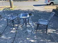 Photo showing the metal patio table and two chairs set arranged on a sidewalk with shadows from nearby trees.