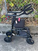Side view of Nexus folding assistance walker showing frame, wheels, padded arm supports with red end caps, and storage pouch beneath seat.