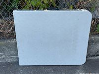 Folded white plastic folding table with rounded corner, leaning against a chain link fence outdoors.