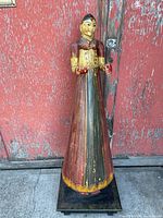Full view of the tall painted wooden statue against a red textured background, showing the entire figure and base.