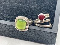 Two modernist style rings, one with a square green stone and one with a round red stone, resting on black display case.