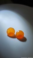 Two natural mandarin orange carnelian gemstones placed on a white surface, shown in soft lighting highlighting their translucency and color.