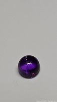 Photo of single deep purple Brazilian amethyst round cabochon gemstone on plain background.