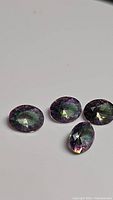 Four oval cut rainbow purple mystic topaz gemstones displayed together on a white background showing color variation and cut.