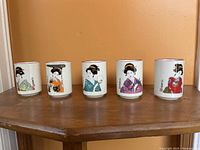 Five ceramic cups lined up, each featuring a different colored Geisha character design on the exterior.