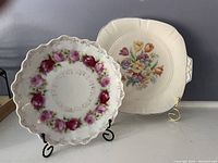 Photo of two antique decorative plates displayed on stands against a gray background