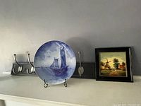 Photo showing a hand painted blue and white tile on stand, an antique framed Dutch plate with windmill scene, and three metal wall hooks in a row.