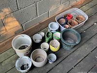 Various garden pots and planters of different materials and sizes arranged on a wooden deck against a concrete wall.