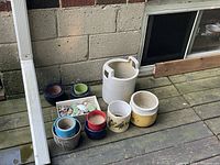 View of 8 garden pots of various sizes and colors along with a small box of decorative stones and ceramic fragments on wooden deck against cinder block wall.