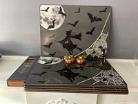 Photo shows 4 Halloween-themed William Sonoma placemats with bats, witch and spider web design, 2 small orange pumpkin candle holders, a brass candlestick holder, and a box of tiny taper candles arranged on a white surface against a neutral background.