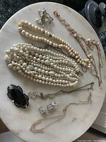 Overview of multiple jewelry pieces on white round surface showing various pearl and bead necklaces, antique brooch, chain necklaces, and clip-on earrings.