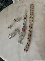Photo showing three pairs of silver-tone stud earrings with clear stones, a multi-stone bracelet with various colored stones, and a long beaded necklace with red beads and pearls all laid out on a white marble surface.