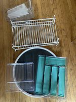 Multiple kitchen organization items on a wooden floor: white wire cupboard shelf rack, large black and white lazy Susan, clear and teal plastic utensil drawer dividers, and a metal basket with plastic handle.