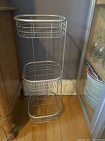 Tall metal wire storage rack with three oval baskets, placed on floor against a wall near wooden furniture and shelf with glassware and plates.