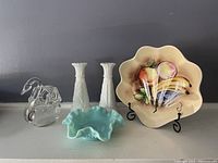Five decorative glass and milk glass items: a clear glass swan ornament, fruit-decorated milk glass plate with crazing on back, two white milk glass candlesticks with embossed pattern, and a blue ruffled-edge glass dish.