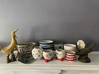 Wide shot of all items on mantel including metal bird doorstop, floral and patterned ceramic planters, novelty egg-shaped planters with faces, floral vase, striped cup and decorative dish.