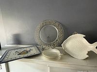 Six cork placemats stacked, rectangular plate with scenic design, round glass plate with gold coral pattern, and large white ceramic fish plate on gold stand