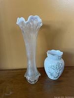 Photo of two antique vases placed side by side on a wooden surface against a beige wall. The left vase is tall, clear milk glass with fluted and ruffled top and vertical texture. The right vase is shorter white stoneware with a green floral design and ruffled opening.