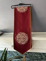 Dark red table runner with beige Chinese New Year motifs and yellow trim hanging on a black hanger