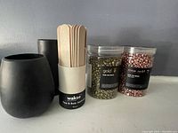 Photo showing two clear containers with metallic gold and rose gold wax beads, a black holder container with wooden applicator sticks, and two black matte wax containers.